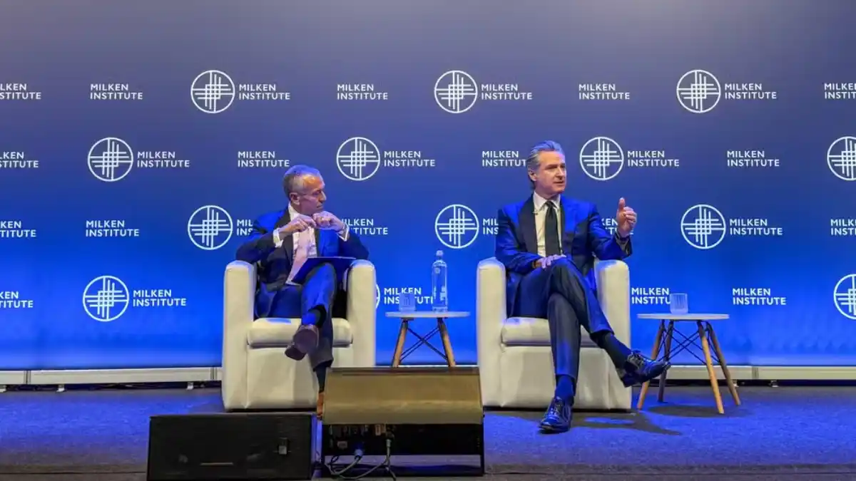 O governador da Califórnia, Gavin Newsom (à direita), sentado no palco do Milken Institute, discursando ao lado de um entrevistador, destacando o Brasil como parceiro.