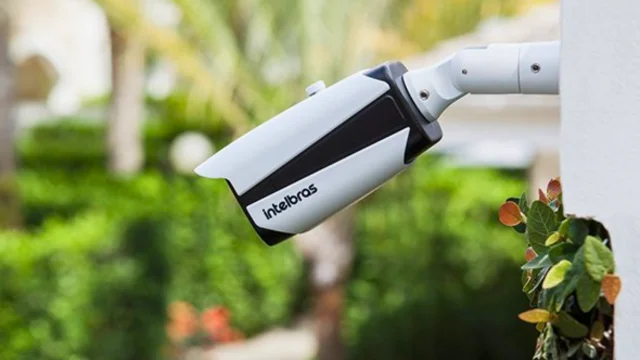 Câmera de segurança branca da Intelbras instalada em uma parede externa com folhagens ao fundo, ilustrando a tecnologia de monitoramento e a análise Intelbras BBI.