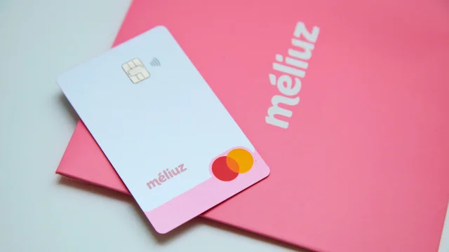 Cartão de crédito físico do Méliuz na cor branca com logotipo rosa, posicionado sobre um envelope de boas-vindas rosa vibrante com a marca Méliuz escrita em branco, ilustrando o serviço de méliuz bitcoin yield.