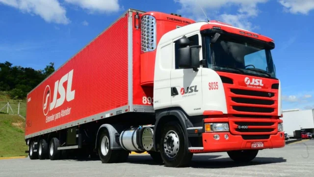 Caminhão baú da JSL pintado em vermelho e branco, com o logotipo da empresa e o slogan "Entender para Atender" na lateral, estacionado em um pátio sob céu azul, representando a frota da jsl safra.