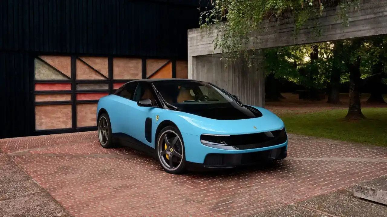 O novo superesportivo elétrico Ferrari Luce em tom azul claro estacionado em um pátio de design moderno. O veículo apresenta um teto preto contrastante, faróis em fendas horizontais e o icônico logotipo da Ferrari no para-lama. Ao fundo, uma garagem com painéis coloridos e uma área arborizada completam o cenário de luxo.
