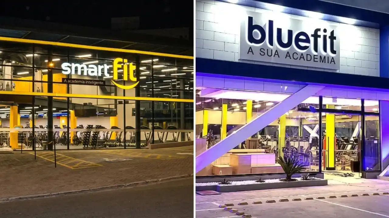 Fachadas de duas academias modernas lado a lado durante o entardecer. À esquerda, uma unidade da SmartFit com sua icônica identidade visual em preto e amarelo e grandes janelas de vidro. À direita, uma unidade da BlueFit com fachada em tons de azul e branco e iluminação em LED. Entre as duas, uma projeção digital exibe um gráfico comparativo de margens Ebitda, onde a curva da SmartFit sobe enquanto a da BlueFit apresenta oscilação e queda.
