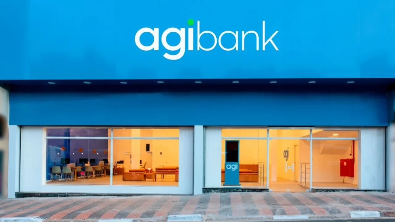 Vista de frente da fachada de uma agência conceito do Agibank (Smart Hub). A parte superior é composta por um grande painel azul escuro com o logotipo "agibank" em letras brancas e horizontais, destacando-se um ponto verde sobre a letra "i". A parte inferior é formada por amplas vitrines de vidro transparente que revelam um interior iluminado e limpo, equipado com mesas de madeira clara, computadores e cadeiras de escritório à esquerda, e uma área de circulação com um totem digital azul vertical escrito "agi" à direita. A entrada fica ao nível da rua, em frente a uma calçada de blocos intertravados nas cores cinza e vermelho.