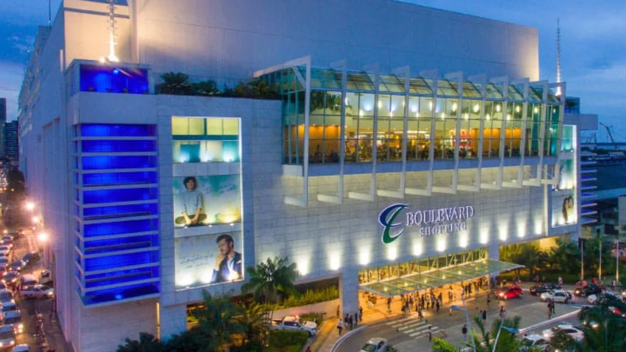 Fotografia em plano aberto e ângulo levemente elevado da fachada externa principal do Boulevard Shopping iluminada ao anoitecer. O edifício de arquitetura moderna exibe uma ampla estrutura de vidro translúcido no andar superior e um letreiro metálico centralizado com o nome "BOULEVARD SHOPPING" ao lado de um logotipo cursivo verde e azul. Na lateral esquerda do prédio, colunas verticais contam com iluminação decorativa em neon azul-brilhante acima de painéis publicitários iluminados. Na parte inferior, observa-se o fluxo de carros na via e pedestres movimentando a entrada do centro comercial.