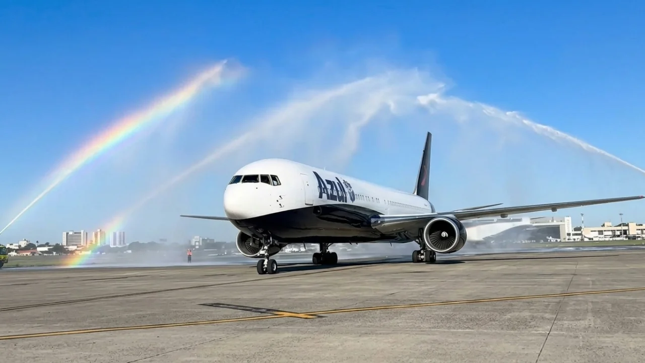 Fotografia em plano aberto de uma aeronave de passageiros de fuselagem larga da Azul Linhas Aéreas na pista de um aeroporto, com fuselagem preta e branca e o logotipo "Azul" azul no nariz e o pássaro rosa no estabilizador vertical. O avião está sendo batizado por dois jatos de água de caminhões de bombeiros do aeroporto sob um céu azul claro e ensolarado. Um arco-íris duplo é visível na névoa da água à esquerda. Prédios do aeroporto e a cidade estão ao fundo.