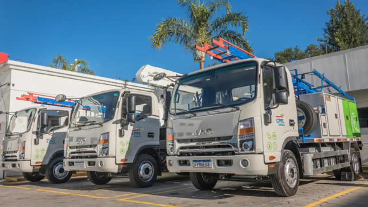 Três caminhões utilitários de serviço brancos da marca JAC, com o logotipo colorido da CPFL Energia estampado nas portas e baús operacionais com faixas verdes na lateral. Os veículos estão estacionados em linha sob um céu azul claro, exibindo escadas laranjas e azuis presas aos tetos. Uma palmeira é visível ao fundo.