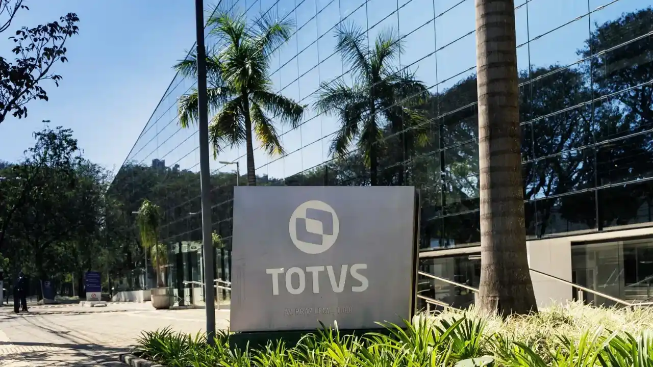 Fotografia em plano médio da entrada principal da sede da Totvs em um dia ensolarado. Em primeiro plano, destaca-se uma placa ou totem cinza com o logotipo circular da empresa e a inscrição em letras brancas "TOTVS AV. BRAZ LEME, 1000", posicionado sobre uma área de paisagismo com vegetação rasteira e gramado verde. Ao fundo, ergue-se o grande edifício comercial com fachada inteiramente de vidro espelhado, refletindo o céu azul claro e copas de palmeiras altas. No lado esquerdo, divisam-se árvores e uma calçada externa.