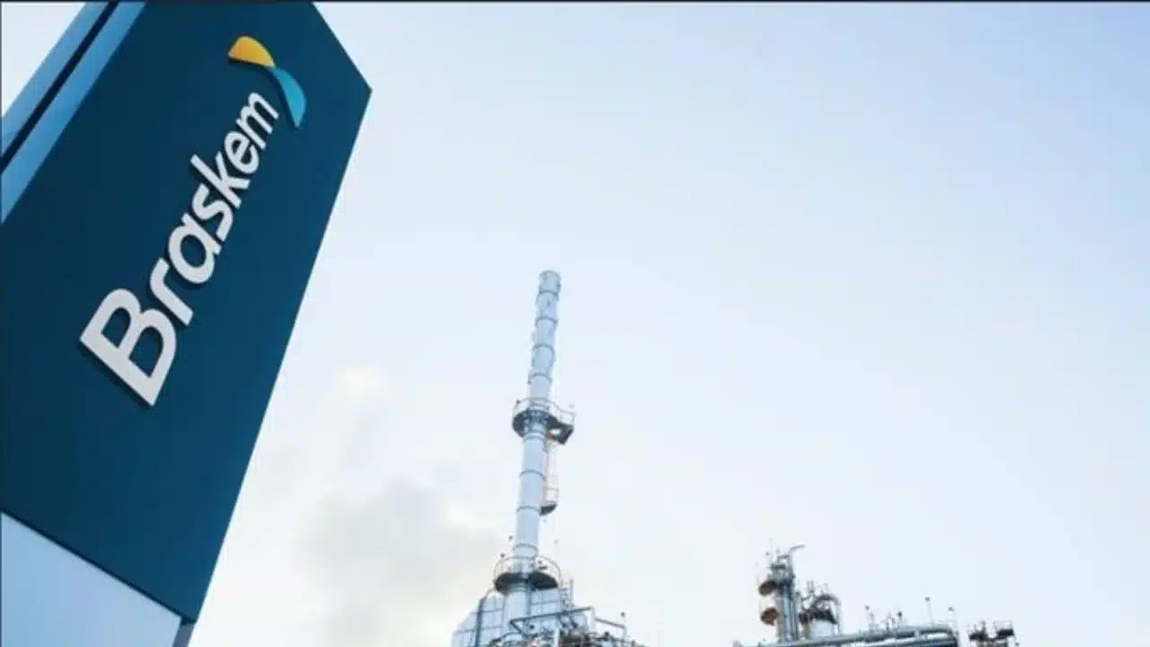 Totem vertical de sinalização corporativa em tom azul-escuro exibindo a palavra "Braskem" em letras brancas com o símbolo gráfico amarelo e ciano no topo. Ao fundo, sob céu limpo de tom azul claro, ergue-se a estrutura metálica de uma usina petroquímica com torres de fracionamento e chaminé industrial.