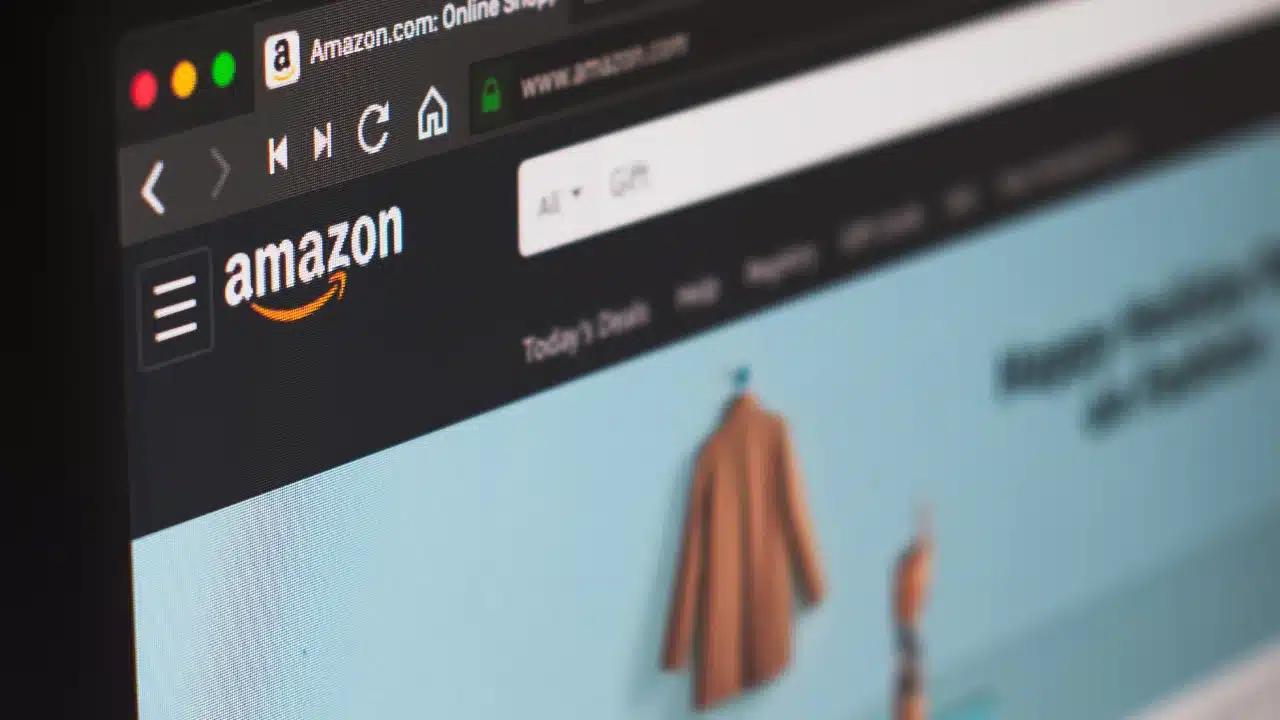 Plano detalhado da tela de um computador portátil exibindo a barra superior do site oficial da Amazon em fundo escuro, destacando o logotipo branco da Amazon com a seta amarela estilizada, a barra de pesquisa e menus da loja online sob navegação do navegador.