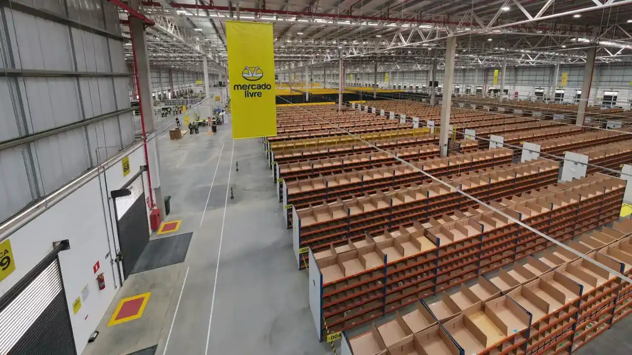 Fotografia em plano geral e ângulo superior (vista aérea interna) de um amplo galpão de centro de distribuição (fulfillment) do Mercado Livre. À direita, organizam-se extensas e simétricas fileiras de estantes metálicas azuis com nichos organizadores de papelão pardo vazios. À esquerda, estende-se um corredor de concreto cinza com marcações de segurança em amarelo e vermelho no piso e portas de docas industriais numeradas como "DOCA 68" e "DOCA 69". No centro, suspenso na alta estrutura metálica do teto, destaca-se um grande banner vertical amarelo com o logotipo oficial do aperto de mãos e a inscrição "mercado livre" escrita em preto. Ao fundo sob luzes industriais, a imensidão do complexo logístico continua com algumas silhuetas de pessoas ao longe.