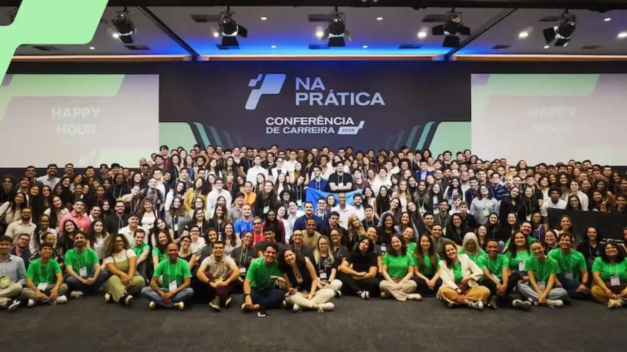 Fotografia em plano aberto e perspectiva frontal de um grande grupo de jovens profissionais e estudantes reunidos no auditório da Conferência de Carreira do Na Prática. No centro, centenas de participantes estão posicionados juntos para uma foto coletiva, muitos deles sentados no chão à frente e outros em pé ao fundo, vestindo camisetas verdes oficiais da organização. Ao fundo, o grande painel digital do palco exibe a identidade visual preta e verde com o logotipo do "Na Prática" e o letreiro centralizado "Conferência de Carreira 2026", ladeado por projeções laterais com a inscrição "Happy Hour". A iluminação cênica do teto destaca a escala e a grande adesão ao evento de empregabilidade.