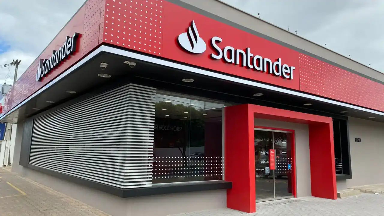 Fotografia em ângulo oblíquo da fachada externa de uma agência bancária do Santander Brasil em dia de céu parcialmente nublado. A estrutura corporativa possui painéis vermelhos e cinzas, com o logotipo da marca "Santander" em letras brancas acompanhado do ícone de chama na cobertura. A entrada principal conta com um pórtico vermelho em destaque e portas de vidro espelhado.