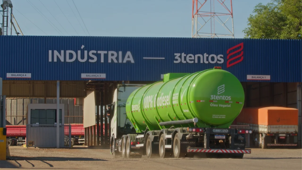 Caminhão-tanque verde com a marca "3tentos Óleo Vegetal" e a indicação "100% Biodiesel" trafegando em pátio fabril ao entrar na balança de pesagem sob uma galeria industrial azul marcada com a escrita "INDÚSTRIA — 3tentos".