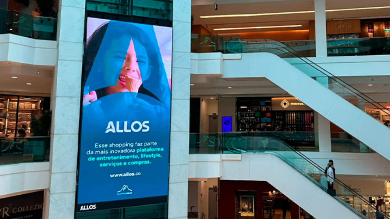 Painel publicitário vertical com o logotipo da Allos no interior de um shopping center moderno com escadas rolantes.