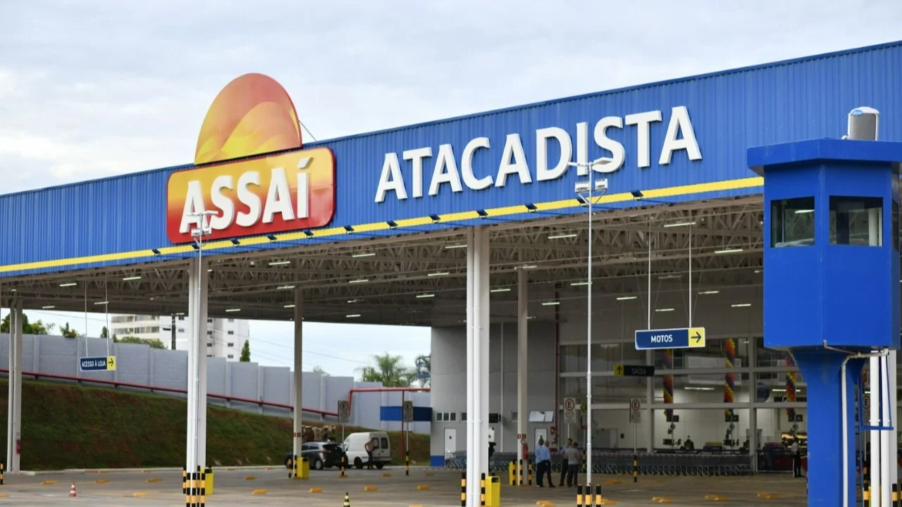 Fachada ampla de unidade do Assaí Atacadista com grande logotipo da marca em amarelo e laranja sobre estrutura azul, área coberta para veículos sustentada por colunas brancas e guarita elevada à direita.