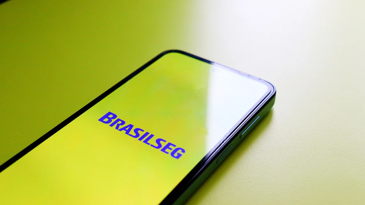 Visão aproximada em ângulo diagonal de um smartphone preto sobre uma superfície clara. A tela do aparelho está acesa em tom amarelo-esverdeado reluzente, exibindo ao centro o logotipo da Brasilseg — empresa de seguros da BB Seguridade — escrito com a palavra "BRASILSEG" em letras maiúsculas azuis.