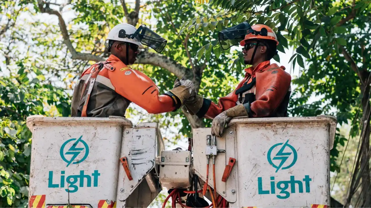 Eletricistas da Light em cesto aéreo se cumprimentando diante do logotipo da concessionária de energia.