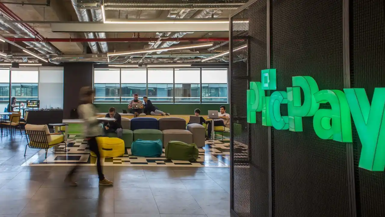 Logotipo do PicPay em verde luminoso na entrada do escritório corporativo com colaboradores ao fundo.