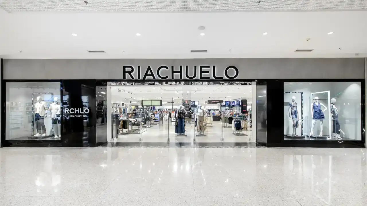 Fachada ampla de uma loja de departamento da Riachuelo em shopping center, exibindo o letreiro preto "RIACHUELO" em destaque no centro superior, vitrines iluminadas com manequins de vestuário feminino e masculino e reflexo no piso claro de porcelanato.