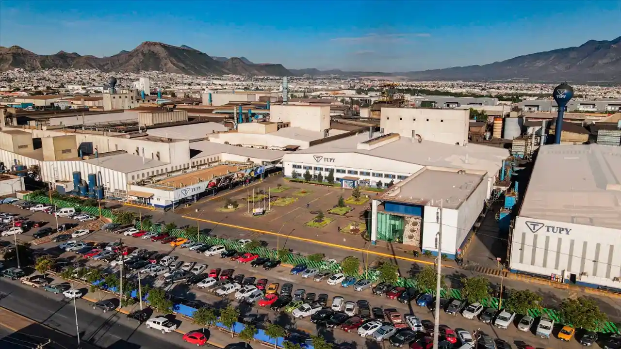Vista aérea panorâmica de grande parque industrial da Tupy com galpões operacionais brancos e beges, logotipo oficial estampado na fachada, pátio lotado de veículos e relevo montanhoso ao fundo sob céu claro.