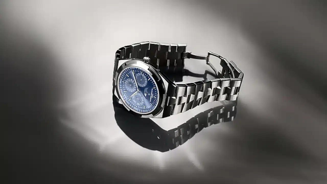 Relógio esportivo de luxo em aço inoxidável com pulseira integrada e mostrador azul, fotografado sobre uma superfície refletiva. A iluminação destaca os acabamentos polidos da caixa e da pulseira, criando reflexos ao redor da peça