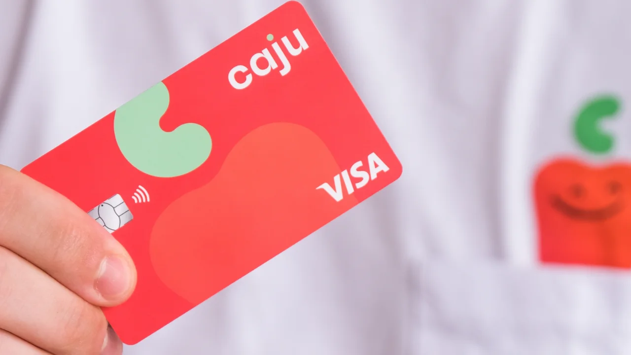 Mão segurando um cartão de benefícios corporativos Caju da bandeira Visa, de cor vermelha com o logotipo em destaque, em frente a uma camisa branca com a estampa da marca Caju.