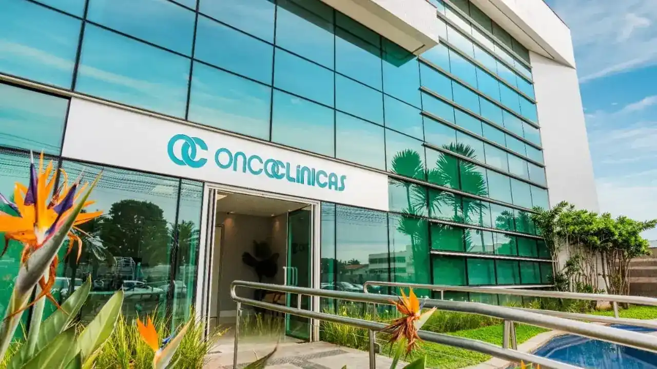 Fachada moderna de vidro espelhado de uma unidade de saúde com o logotipo e nome Oncoclínicas em destaque sobre a entrada principal e jardim tropical ao redor.