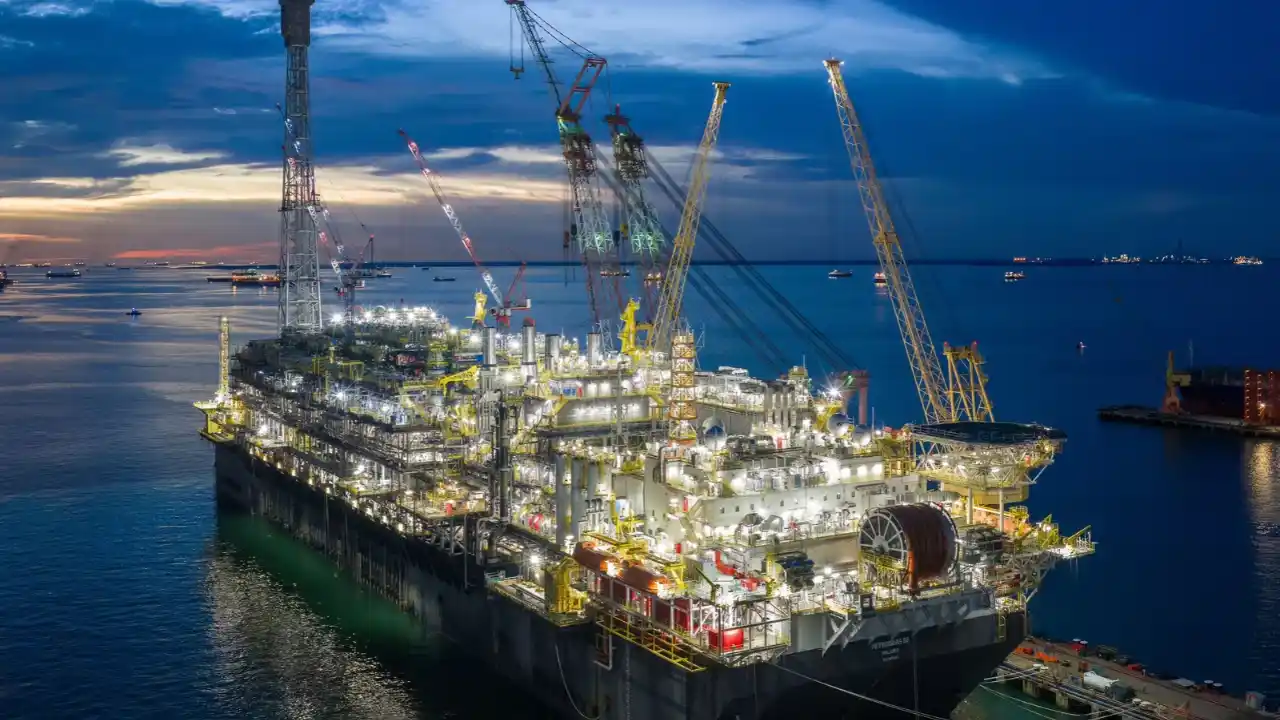 Vista aérea ao anoitecer da plataforma de petróleo P-82 iluminada em águas calmas, mostrando guindastes, módulos industriais e a estrutura do FPSO ancorado próximo ao porto.