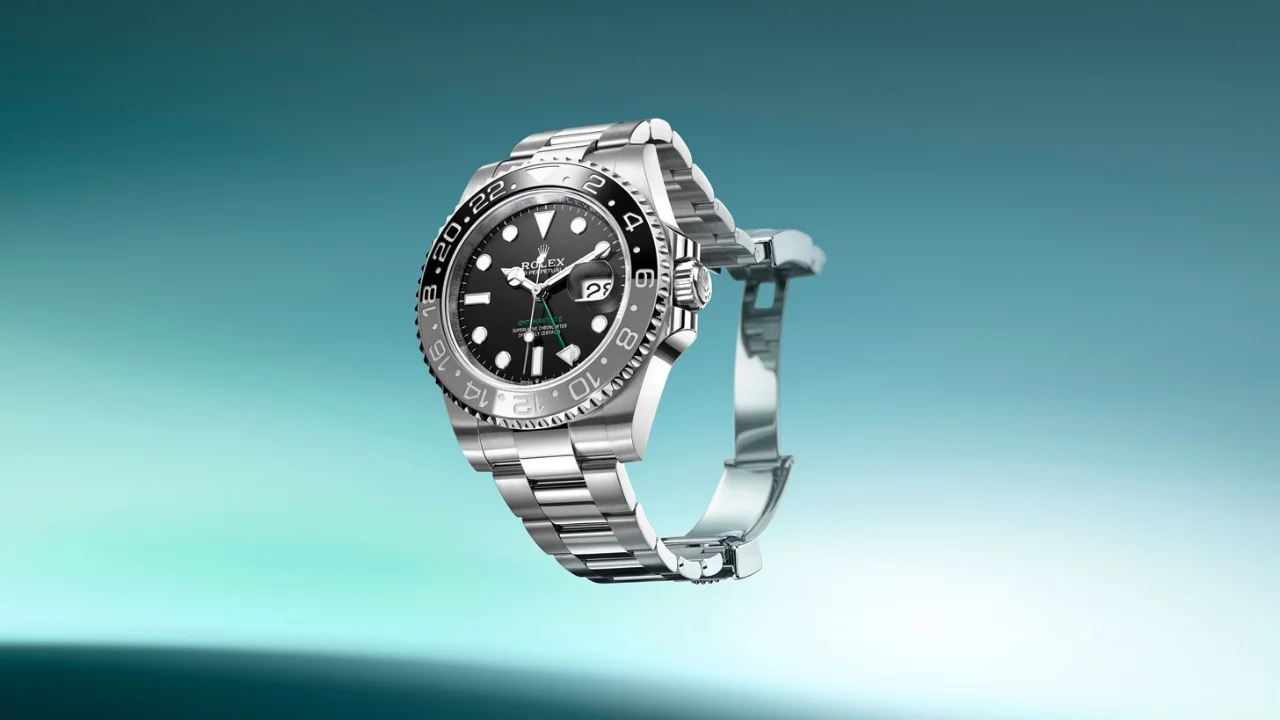 Relógio de luxo Rolex Oyster Perpetual GMT-Master II em aço inoxidável, com mostrador preto, bisel giratório bidirecional em cerâmica preta e cinza e pulseira metálica sobre fundo em tom azul-esverdeado.